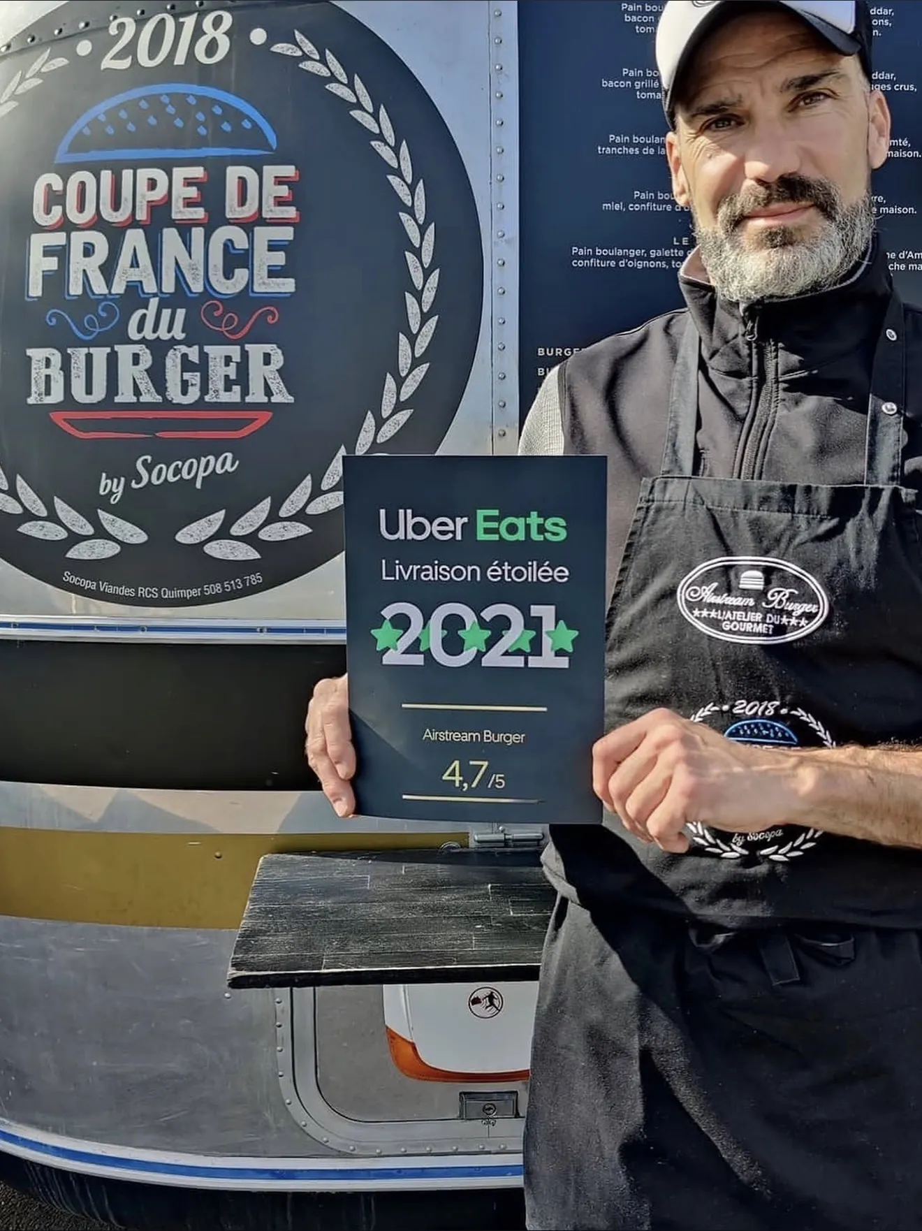 Brice devant l'Airstream avec le diplôme Uber Eats Livraison étoilée 2021 et le macaron Coupe de France du Burger 2018